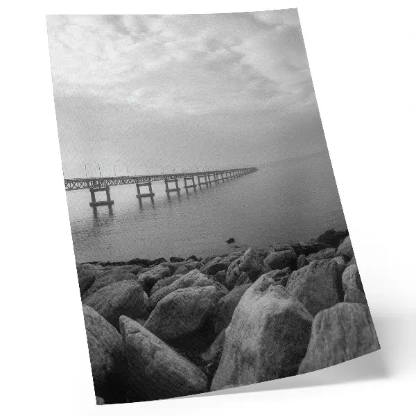 Nowoczesna grafika na płótnie spokojny pejzaż z mostem na kamienistej plaży przy szarym niebie